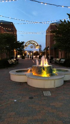 a fountain in the middle of a brick walkway with lights strung over it and people walking by