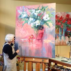 an older woman standing in front of a painting on easel holding a flower vase