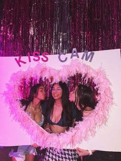 three women standing in front of a sign that says kiss cam with pink feathers on it