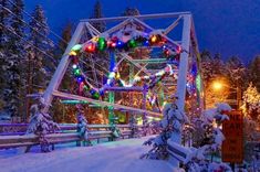 a bridge with christmas lights on it in the middle of snow covered trees and evergreens