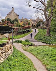 two people riding bikes down a road in front of some stone buildings and trees with green grass on both sides