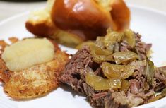 a white plate topped with meat and potatoes next to breaded buns on a table