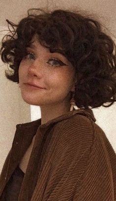 a close up of a person wearing a brown jacket and smiling at the camera with curly hair