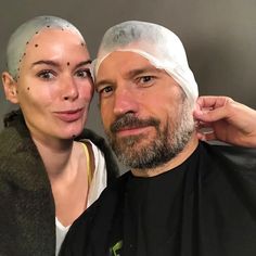 a man and woman pose for a selfie in front of a gray wall with black dots on their foreheads