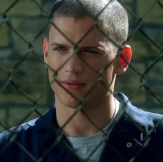 a young man behind a chain link fence looking at the camera with an evil look on his face