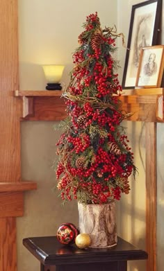 a christmas tree with red berries and pine cones on a small table in front of a fireplace