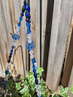 a blue glass beaded necklace hanging from a plant in front of a wooden fence