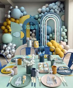 a table set with plates, cups and silverware in front of a wall decorated with balloons