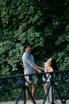 a man and woman holding hands while standing on a bridge in front of some trees