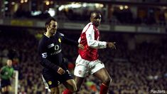 two men playing soccer on a field with people watching from the stands in the background