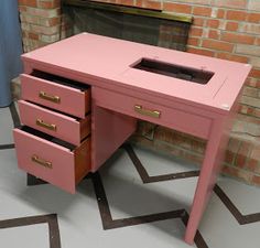 a pink desk with three drawers in front of a fire place and brick wall behind it