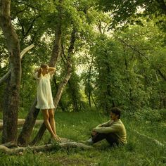 two people are sitting in the grass near some trees and one person is leaning up against a tree