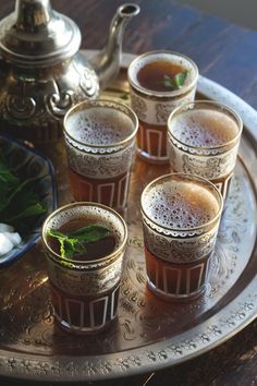 four shot glasses filled with liquid and mint on a silver tray next to a teapot