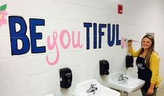 a woman standing next to two sinks in a bathroom with the words be you tiful painted on the wall