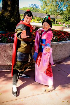 two people dressed up in costumes posing for a photo on the sidewalk near a flower garden