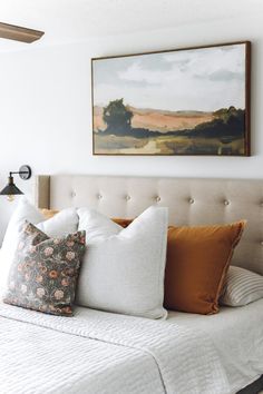 a bed with white linens and orange pillows in a small room next to a painting on the wall