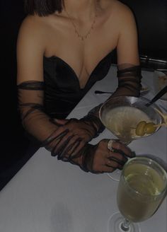 a woman wearing gloves sitting at a table with two glasses and a drink in front of her