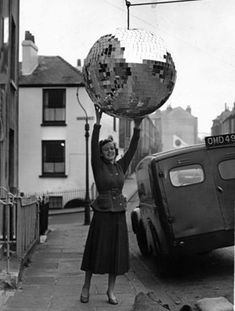 a woman holding up a disco ball in front of a car on the side of the road