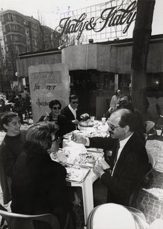 a group of people sitting around a table eating food and drinking wine in front of a sign