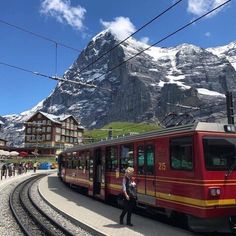 a red train traveling down tracks next to snow covered mountain range in the distance with people walking on it