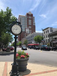 a clock sitting on the side of a road next to a planter filled with flowers