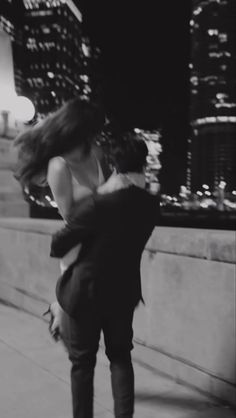 a man and woman hugging on the sidewalk in front of a cityscape at night