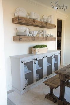 a dining room table and some shelves with dishes on top of them, in front of a white wall