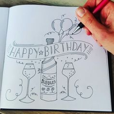 a hand is holding a pen and writing happy birthday on a book with wine glasses