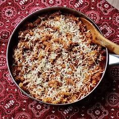 a skillet filled with pasta and cheese on top of a red table cloth next to a wooden spoon