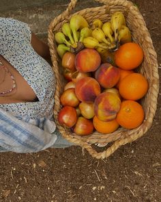 a woman sitting on the ground next to a basket full of peaches and bananas