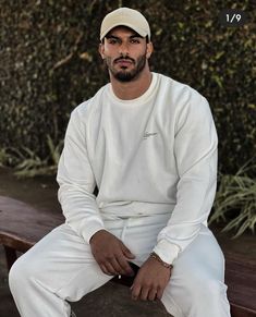 a man sitting on top of a wooden bench wearing a white shirt and pants with his hands in his pockets