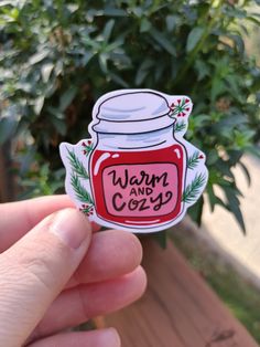 a hand holding up a sticker with the words warm and cozy in front of a potted plant