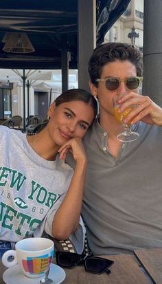 a man and woman sitting at a table with drinks in front of them, both holding their arms around each other