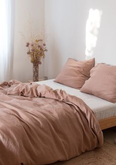 a bed with pink sheets and pillows in a white room next to a vase filled with flowers