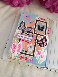 an open notebook with butterflies and flowers on the cover, next to some pink flowers