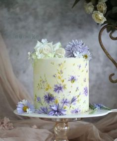 a white cake with purple and yellow flowers sitting on top of a glass stand next to a flower bouquet