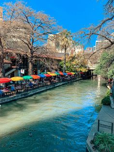 people sitting at tables on the side of a river with umbrellas over their heads