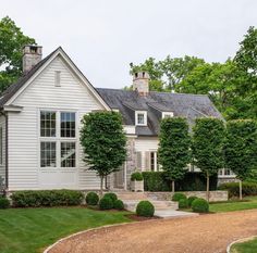 a large white house surrounded by lush green trees and bushes in front of it, with a gravel driveway leading to the front door