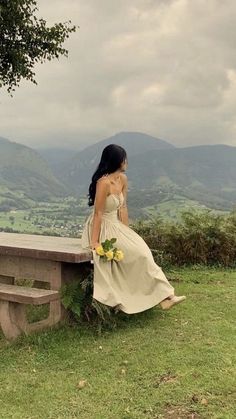 a woman in a dress sitting on a bench looking out at the valley and mountains