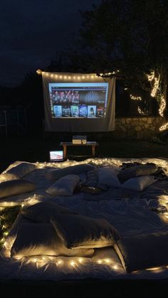 an outdoor movie screen is lit up at night with lights on the ground and pillows all around it