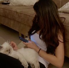 a woman sitting on the floor holding a cat and using a cell phone to play with it