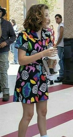 a woman in a colorful dress is holding a cup and standing on the sidewalk with other people