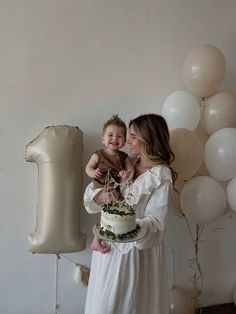 a woman holding a small child in front of balloons and a cake with the number one on it