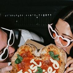 a man and woman holding up a heart shaped pizza in front of their face with the word love spelled on it