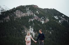 a pregnant couple holding hands while standing in front of a mountain