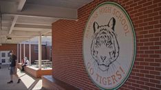 two people are walking past a sign for the high school tigers on a brick wall