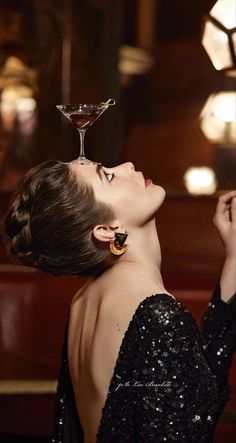 a woman in a black dress is holding a martini glass up to her face and looking upward