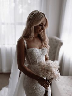 a woman in a wedding dress is holding a bouquet and looking at the camera while standing next to a bed