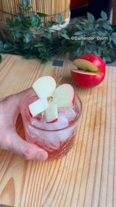 a person holding an apple on top of a wooden table next to a glass filled with ice