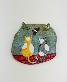 two cats sitting on top of a red and blue rock with butterflies flying over it